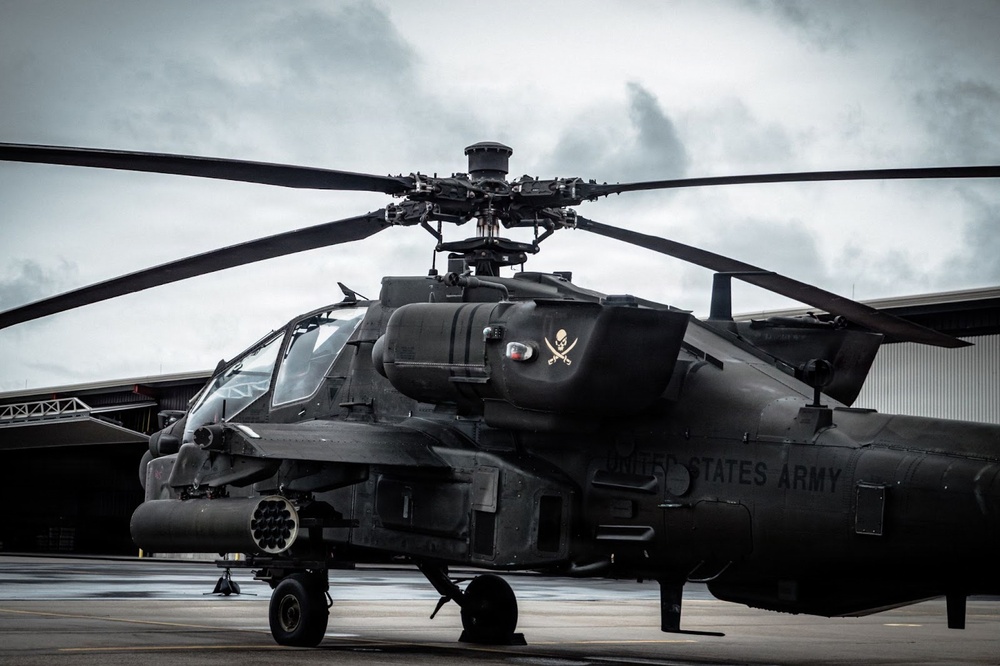 A United States Army AH-64 Apache attack helicopter is parked on a tarmac in front of a hangar under overcast skies. The aircraft is painted in a dark matte finish and displays a skull-and-crossbones insignia on its fuselage. The helicopter is shown in a static ground position with rotor blades extended.