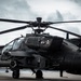 A United States Army AH-64 Apache attack helicopter is parked on a tarmac in front of a hangar under overcast skies. The aircraft is painted in a dark matte finish and displays a skull-and-crossbones insignia on its fuselage. The helicopter is shown in a static ground position with rotor blades extended.