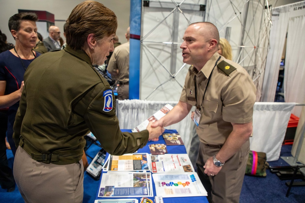 Two military personnel are shaking hands at what appears to be a military expo or career fair. The individual on the left wears a dark green Army uniform with an Airborne patch (173rd Airborne Brigade), while the individual on the right wears a tan/khaki uniform with a Major rank insignia. A table covered with informational brochures and pamphlets is visible between them.