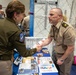 Two military personnel are shaking hands at what appears to be a military expo or career fair. The individual on the left wears a dark green Army uniform with an Airborne patch (173rd Airborne Brigade), while the individual on the right wears a tan/khaki uniform with a Major rank insignia. A table covered with informational brochures and pamphlets is visible between them.