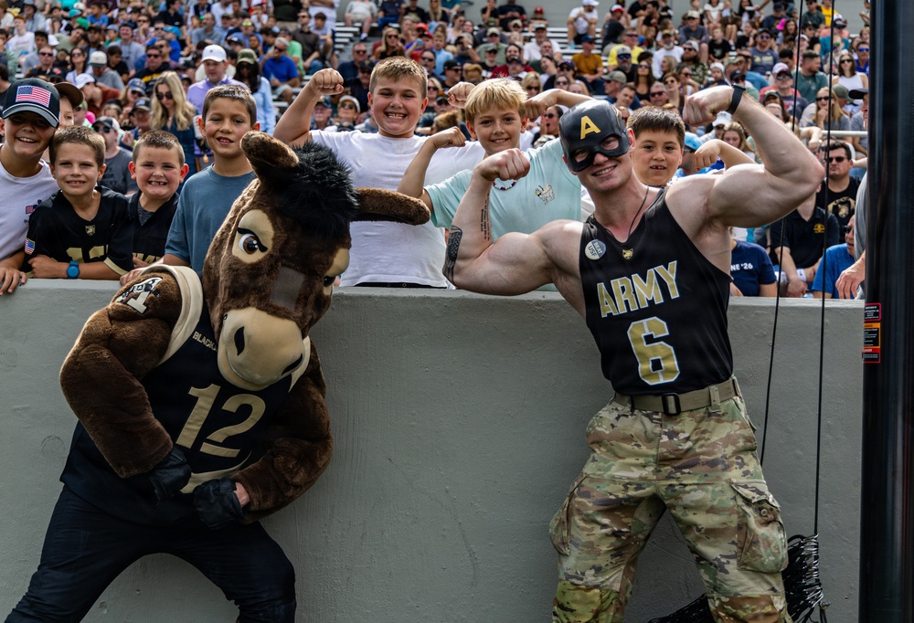 A lively scene at a US Army (West Point) football game featuring the team's mule mascot wearing jersey number 12 and a muscular man in an Army jersey number 6 wearing a Captain America-style mask, both posing with flexing biceps. A crowd of enthusiastic children and adults lean over a stadium wall behind them, many also flexing their arms in imitation. The atmosphere is festive and energetic, consistent with a college football game event.