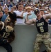 A lively scene at a US Army (West Point) football game featuring the team's mule mascot wearing jersey number 12 and a muscular man in an Army jersey number 6 wearing a Captain America-style mask, both posing with flexing biceps. A crowd of enthusiastic children and adults lean over a stadium wall behind them, many also flexing their arms in imitation. The atmosphere is festive and energetic, consistent with a college football game event.