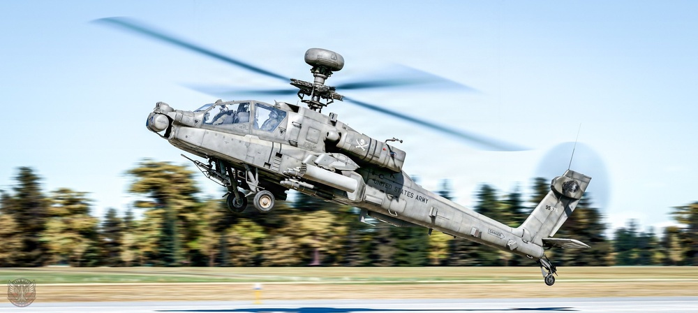 A United States Army AH-64 Apache attack helicopter is captured in dynamic low-level flight, appearing to be taking off or performing a high-speed pass at low altitude. The image uses a panning photography technique, creating motion blur in the background while keeping the aircraft relatively sharp. The helicopter is armed and equipped with a Longbow radar dome mounted above the rotor hub.