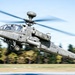 A United States Army AH-64 Apache attack helicopter is captured in dynamic low-level flight, appearing to be taking off or performing a high-speed pass at low altitude. The image uses a panning photography technique, creating motion blur in the background while keeping the aircraft relatively sharp. The helicopter is armed and equipped with a Longbow radar dome mounted above the rotor hub.