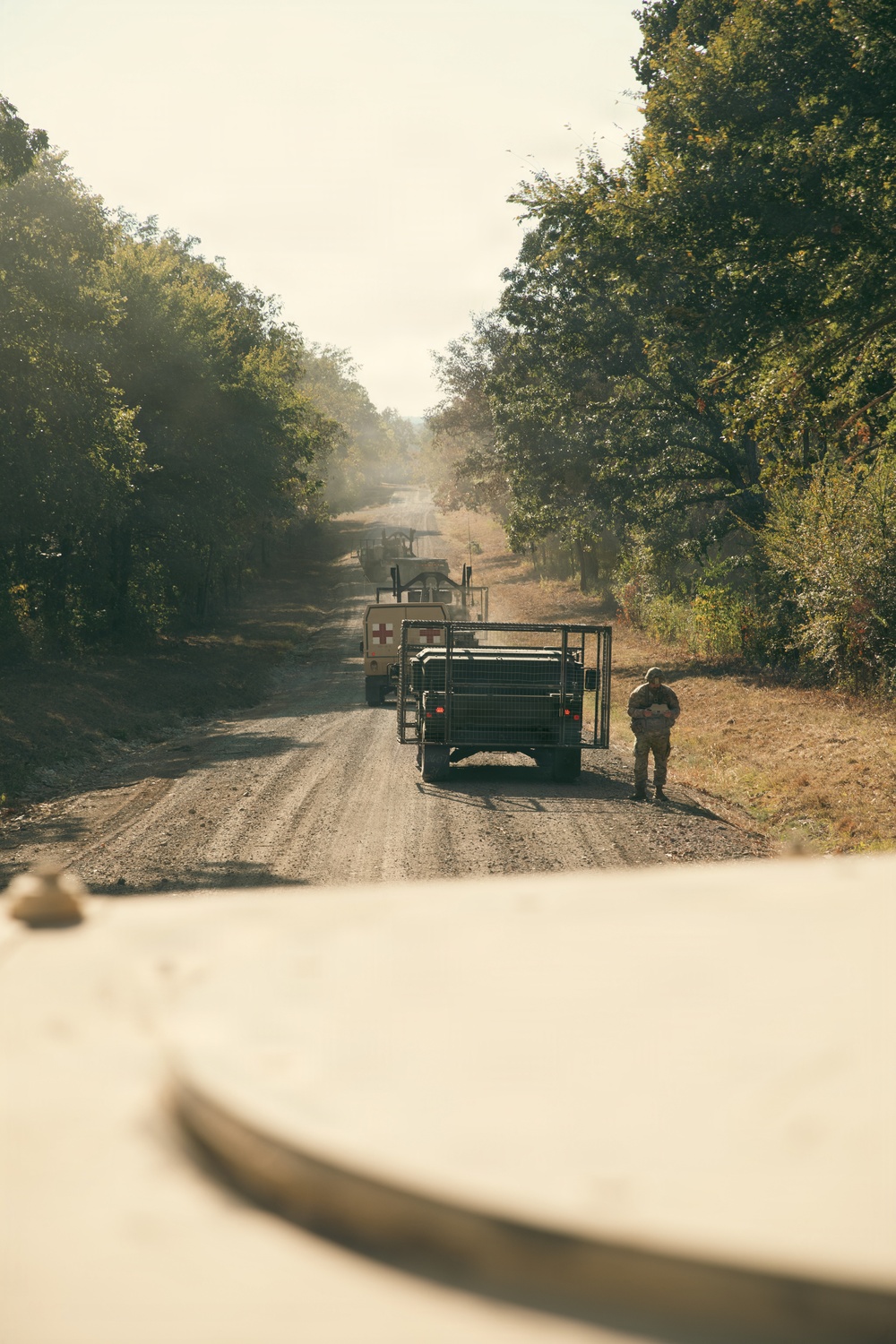 A military convoy is halted on a dusty dirt road flanked by dense trees, photographed from inside a lead vehicle. At least two military trucks are visible, one bearing a Red Cross medical marking, with a soldier standing beside the rear vehicle. The scene appears to depict a temporary convoy stop in a rural or semi-forested area.