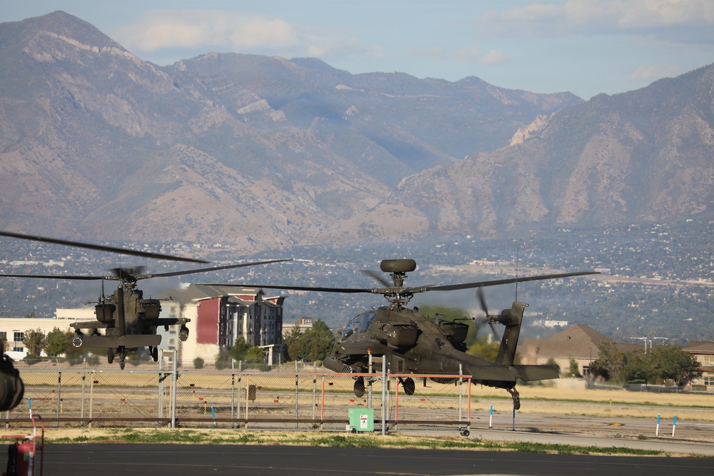 Two military attack helicopters, identifiable as AH-64 Apache variants, are captured in low-altitude flight or hover near what appears to be a military airfield or helipad. The scene is set against a backdrop of a large mountain range, likely the Wasatch Mountains near Salt Lake City, Utah. Urban development and commercial buildings are visible in the middle ground between the helicopters and the mountains.