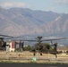 Two military attack helicopters, identifiable as AH-64 Apache variants, are captured in low-altitude flight or hover near what appears to be a military airfield or helipad. The scene is set against a backdrop of a large mountain range, likely the Wasatch Mountains near Salt Lake City, Utah. Urban development and commercial buildings are visible in the middle ground between the helicopters and the mountains.