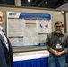 Three researchers stand in front of a scientific poster at what appears to be an academic or medical conference. The poster, sponsored by the Defense Health Agency (DHA) and WRAIR, presents research on 'Multi-omics mechanisms tracking the longitudinal path towards return to action from contact sports-induced TBI.' Two of the three individuals are wearing visible name badges identifying them as 'Nabarun Chakraborty' and 'Swapna Kannan.'