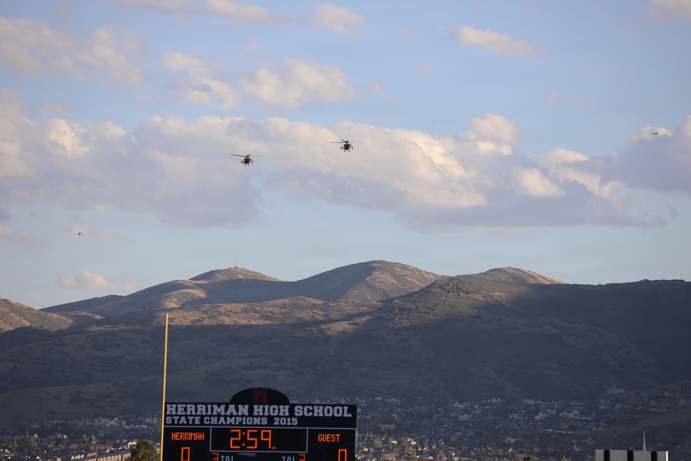 The image shows a football scoreboard for Herriman High School, displaying '2:59' on the game clock with a score of 0-0 between Herriman and Guest. Multiple military helicopters are visible flying in formation over a mountainous landscape. The scene appears to be a ceremonial flyover during a high school football game, with the scoreboard noting 'State Champions 2015'.
