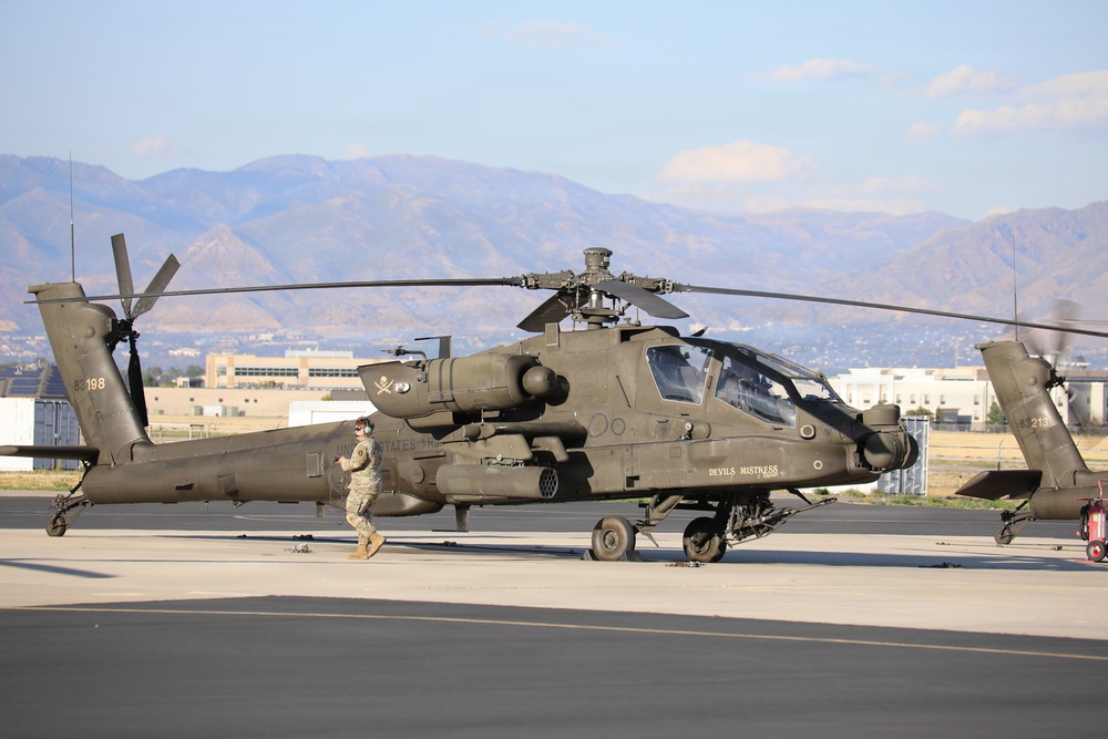 A U.S. Army AH-64 Apache attack helicopter, named 'Devils Mistress,' is parked on a tarmac at a military airfield. A soldier in camouflage uniform is conducting a pre-flight or post-flight inspection walk-around. A second Apache helicopter is partially visible on the right side of the frame.