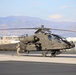 A U.S. Army AH-64 Apache attack helicopter, named 'Devils Mistress,' is parked on a tarmac at a military airfield. A soldier in camouflage uniform is conducting a pre-flight or post-flight inspection walk-around. A second Apache helicopter is partially visible on the right side of the frame.
