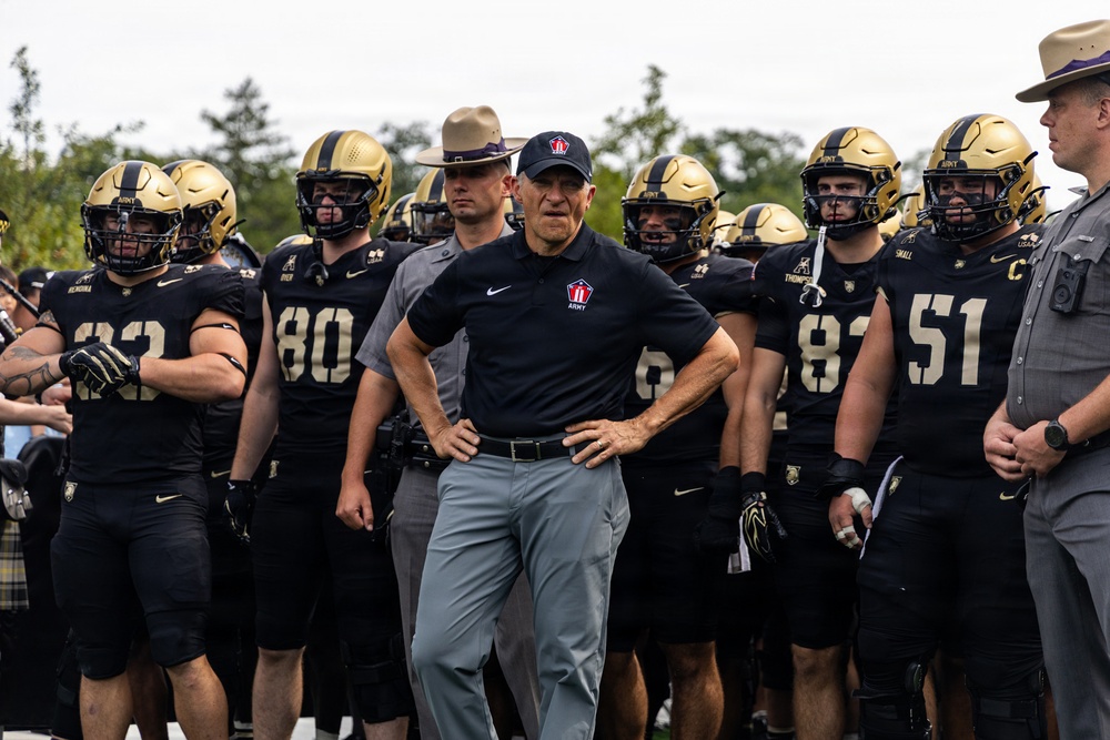 A football coach stands at the center of a group of players wearing black and gold uniforms with 'ARMY' branding, appearing to be preparing to take the field. The coach is dressed in a black Nike polo shirt with an Army logo, gray pants, and a black cap. Two individuals in gray uniforms with campaign/drill sergeant-style hats are also visible flanking the group.