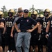 A football coach stands at the center of a group of players wearing black and gold uniforms with 'ARMY' branding, appearing to be preparing to take the field. The coach is dressed in a black Nike polo shirt with an Army logo, gray pants, and a black cap. Two individuals in gray uniforms with campaign/drill sergeant-style hats are also visible flanking the group.