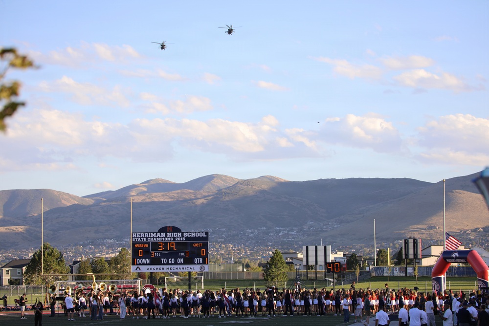 A high school football game or pre-game event at Herriman High School, with two military helicopters performing a flyover above the stadium. The scoreboard identifies the school as 'Herriman High School State Champions 2013' and shows a score of 3:19 with sponsorship by Archer Mechanical. A large crowd, marching band, and cheerleaders are gathered on the field with mountains visible in the background.