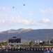 A high school football game or pre-game event at Herriman High School, with two military helicopters performing a flyover above the stadium. The scoreboard identifies the school as 'Herriman High School State Champions 2013' and shows a score of 3:19 with sponsorship by Archer Mechanical. A large crowd, marching band, and cheerleaders are gathered on the field with mountains visible in the background.