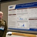 A military service member in Army dress uniform stands beside a scientific research poster at what appears to be an academic or military research conference. The poster is titled 'Evaluating microRNAs as Biomarkers for Traumatic Brain Injury and Blast Overpressure' and is affiliated with the Defense Health Agency (DHA). The presenter's name tag reads 'Bryanna Schermerhorn' and she is associated with WRAIR (Walter Reed Army Institute of Research).