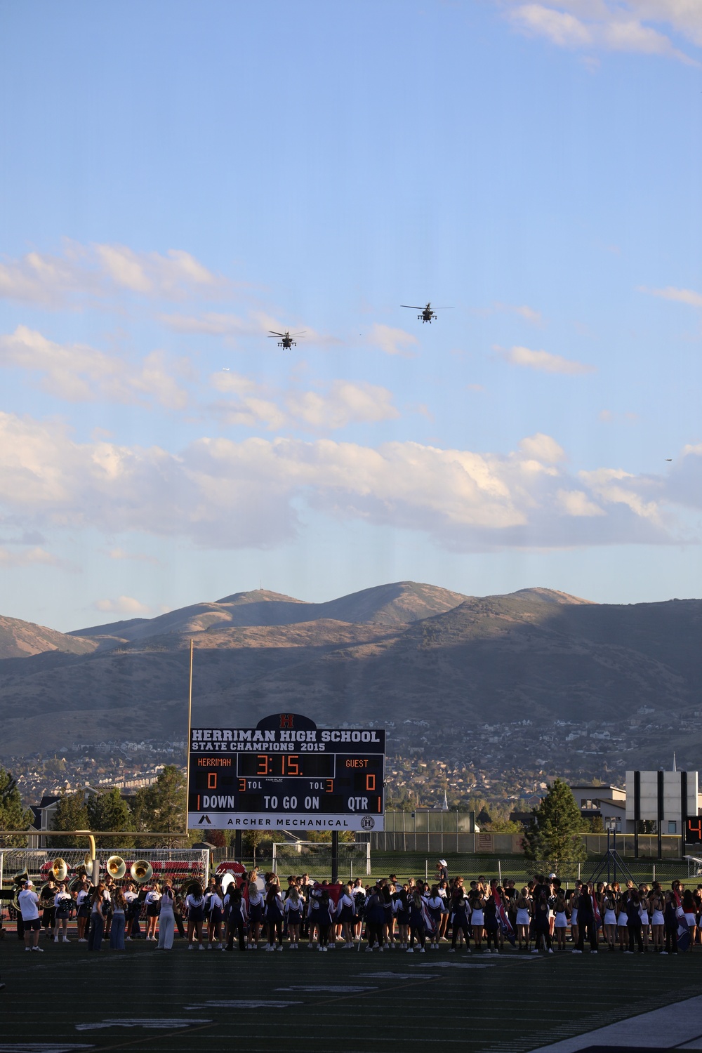 A high school football game at Herriman High School in Utah features two military helicopters performing a flyover above the stadium. The scoreboard displays the game clock at 3:15 in the 1st quarter with both teams at 0, and notes the school's 2015 State Championship. Cheerleaders, band members, and other students are gathered along the sideline watching the aerial display.