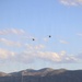 A high school football game at Herriman High School in Utah features two military helicopters performing a flyover above the stadium. The scoreboard displays the game clock at 3:15 in the 1st quarter with both teams at 0, and notes the school's 2015 State Championship. Cheerleaders, band members, and other students are gathered along the sideline watching the aerial display.