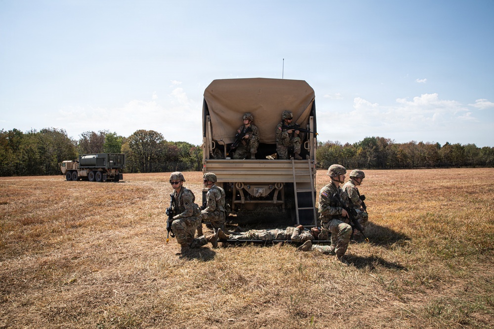Military personnel in camouflage uniforms are conducting what appears to be a field training exercise in an open dry grassland area. Several soldiers are positioned around a tan military truck with a canvas-covered bed, with two soldiers elevated in the truck bed and others kneeling on the ground around a figure lying on a litter/stretcher. Additional military vehicles are visible in the background near a tree line.