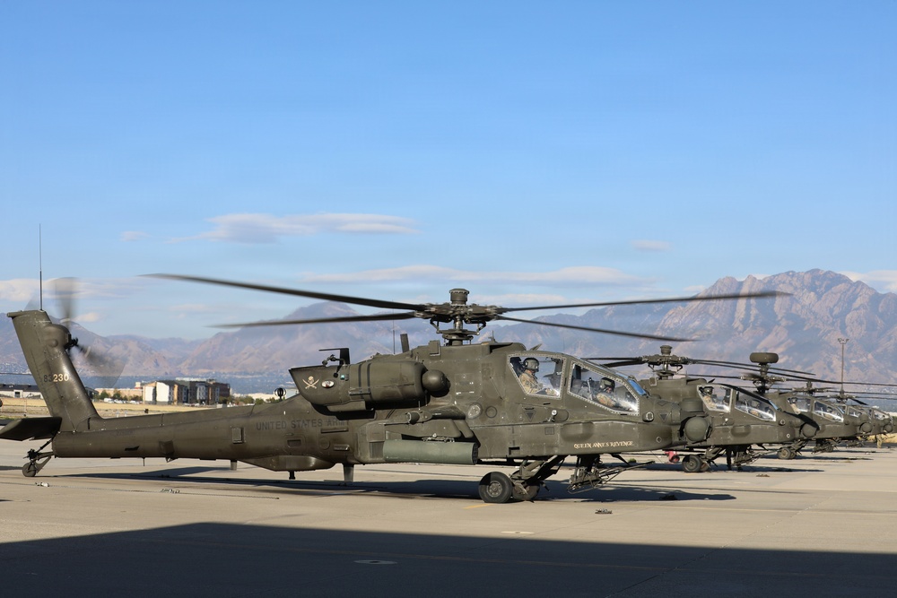 Multiple U.S. Army AH-64 Apache attack helicopters are lined up on a tarmac, with at least one having its rotors spinning. The helicopters are painted in olive drab and bear 'United States Army' markings, with one visibly named 'Queen Anne's Revenge'. Mountains and a partially cloudy blue sky form the backdrop, suggesting a western U.S. military installation.