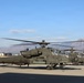 Multiple U.S. Army AH-64 Apache attack helicopters are lined up on a tarmac, with at least one having its rotors spinning. The helicopters are painted in olive drab and bear 'United States Army' markings, with one visibly named 'Queen Anne's Revenge'. Mountains and a partially cloudy blue sky form the backdrop, suggesting a western U.S. military installation.