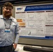 A young man named Joseph Dwyer, identified by his name badge as being from 'The Geneva Foundation,' stands beside a scientific research poster at what appears to be an academic or military medical conference. The poster, sponsored by DHA (Defense Health Agency), presents research on iontophoretic delivery of glyburide for treating traumatic brain injury in swine. The setting appears to be a conference exhibition hall with numbered booth markers visible.