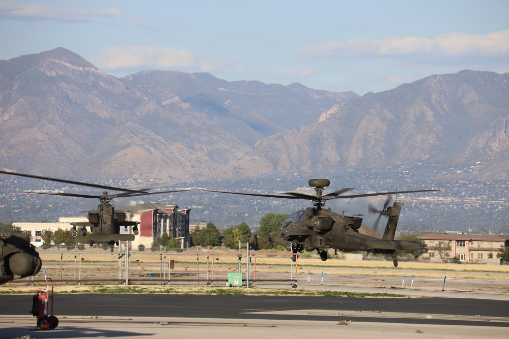 Two military attack helicopters, identifiable as AH-64 Apache variants, are captured in low-altitude flight or hover over a military airfield tarmac. A mountain range rises prominently in the background, with urban development and military installation buildings visible in the mid-ground. The image appears to be taken at a U.S. Army installation, likely in the southwestern United States based on the terrain.