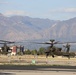 Two military attack helicopters, identifiable as AH-64 Apache variants, are captured in low-altitude flight or hover over a military airfield tarmac. A mountain range rises prominently in the background, with urban development and military installation buildings visible in the mid-ground. The image appears to be taken at a U.S. Army installation, likely in the southwestern United States based on the terrain.