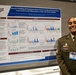 A U.S. Army soldier named Ayana Ferrer (visible on name tag) stands smiling beside a scientific research poster titled 'Kinetic Profiling of Traumatic Brain Injury and Blast Overpressure-Associated Serum Biomarkers in Rat and Swine Models.' The poster is affiliated with the Defense Health Agency (DHA) Research &amp; Development and Walter Reed Army Institute of Research. The setting appears to be an academic or military medical research conference.