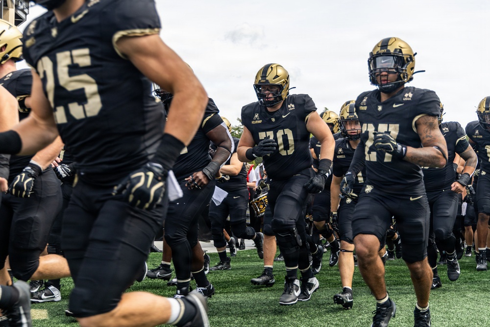 A college football team dressed in black and gold uniforms is running onto the field, likely at the start of a game. The players are wearing black helmets with gold accents and Nike gear, with visible jersey numbers including 25, 70, and 27. The scene captures the energy of a pre-game entrance on a green turf field.