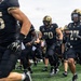 A college football team dressed in black and gold uniforms is running onto the field, likely at the start of a game. The players are wearing black helmets with gold accents and Nike gear, with visible jersey numbers including 25, 70, and 27. The scene captures the energy of a pre-game entrance on a green turf field.