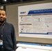A man named Burook Msganaw, as identified by his conference badge, stands beside a scientific research poster at what appears to be an academic or military research conference. The poster is titled 'Dose-Response Transcriptomic Dynamics in a Gyrencephalic Ferret Model of Blast-Induced Traumatic Brain Injury' and features the DHA (Defense Health Agency) logo. The setting is a conference exhibition hall with booth number 2102 visible in the background.