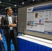 Two individuals are viewing a scientific research poster at what appears to be a military or defense health conference. The poster, displayed under DHA (Defense Health Agency) and WRAIR (Walter Reed Army Institute of Research) branding, presents research titled 'Dysregulation of Early Metabolic Changes in the Lungs and Plasma Following Blast Exposure in Ferret and Mouse Models.' A man in a dark suit appears to be presenting or discussing the poster with a woman in a military tan uniform.