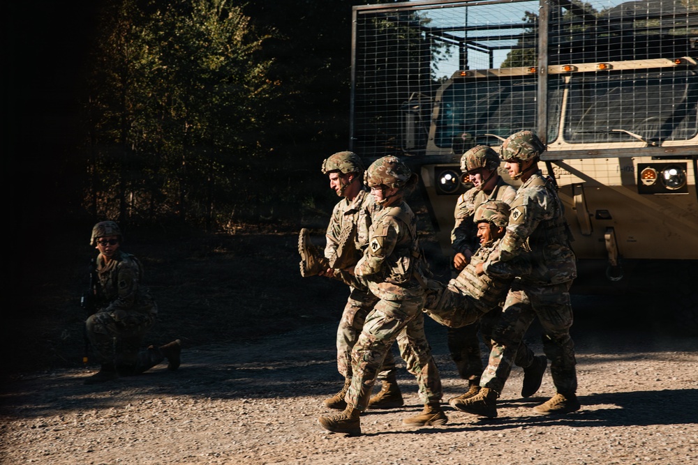 A group of soldiers in US Army camouflage uniforms are conducting what appears to be a casualty evacuation drill or training exercise, with multiple personnel carrying a fellow soldier. A military tactical vehicle (likely an LMTV or similar) is visible in the background, and a lone soldier is kneeling in the shadows to the left. The scene takes place on a gravel surface near a wooded area in what appears to be daytime training conditions.