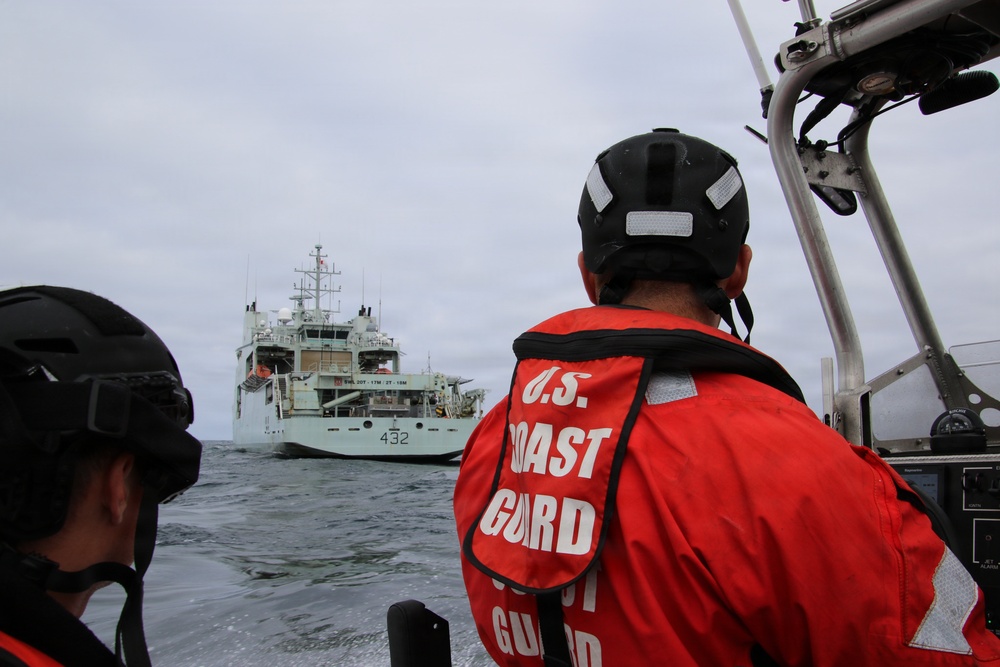 Two U.S. Coast Guard personnel aboard a small patrol vessel are observing a larger gray naval/coast guard ship bearing hull number 432 in open water. The personnel are wearing distinctive orange immersion suits labeled 'U.S. Coast Guard' and black helmets with reflective strips. The scene appears to depict an at-sea rendezvous or approach operation under overcast skies.
