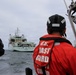 Two U.S. Coast Guard personnel aboard a small patrol vessel are observing a larger gray naval/coast guard ship bearing hull number 432 in open water. The personnel are wearing distinctive orange immersion suits labeled 'U.S. Coast Guard' and black helmets with reflective strips. The scene appears to depict an at-sea rendezvous or approach operation under overcast skies.