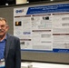 A man named James DeMar, identified by his conference badge, stands in front of a scientific research poster at what appears to be an academic or military research conference. The poster is titled 'Acute Phosphoproteome Changes in the Brain of Ferrets after Blast Exposure' and is affiliated with the Defense Health Agency (DHA). The setting is a conference exhibition hall with numbered booth markers visible in the background.