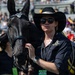 A woman wearing a black cowboy hat, sunglasses, and an Army West Point staff shirt stands beside a black horse with a decorated bridle at what appears to be a stadium event. She is holding the horse's bridle with red-painted fingernails and wearing a smartwatch and a green event credential badge. The background shows stadium bleachers filled with spectators.