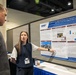 A woman named Megan (visible on her name badge) is presenting a scientific research poster at what appears to be an academic or military health conference. The poster, affiliated with the Defense Health Agency (DHA), is titled 'Comparison of 3 Different Mortar Fire Exposure Environments and Their Acute Neurological Effects.' She is pointing at the poster while explaining its contents to a man in a gray suit.