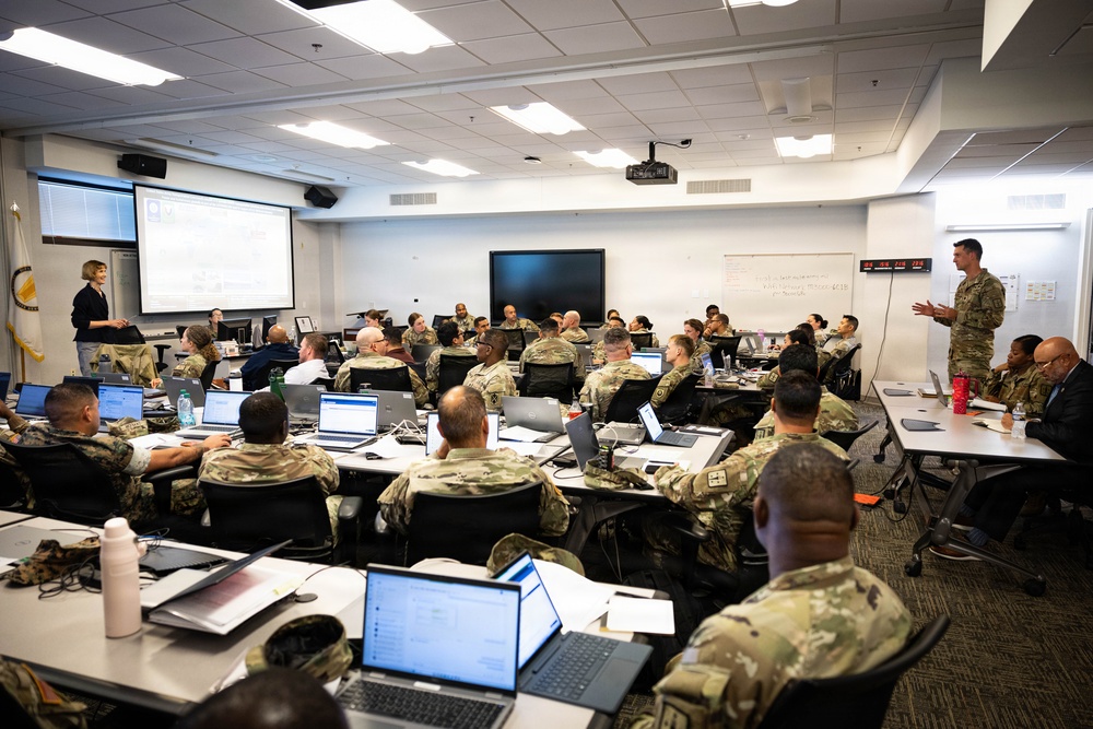 A large military training or briefing session is taking place in a modern classroom or conference room. Numerous personnel in U.S. Army camouflage uniforms (OCP pattern) are seated at tables with open laptops, attending a presentation. Two instructors or briefers are visible at the front of the room, one civilian and one uniformed soldier.