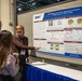A researcher wearing a hijab is presenting a scientific poster titled 'Gene-Epigene Cross-Tissue Signatures in Response to Blast Injury in Mouse Model' at what appears to be an academic or military research conference. She is pointing at the poster while engaging with two attendees, one of whom is holding a coffee cup. The poster is affiliated with DHA (Defense Health Agency) and covers topics including Traumatic Brain Injury, miRNA/mRNA Transcriptomics, and Cross-Tissue Comparison.