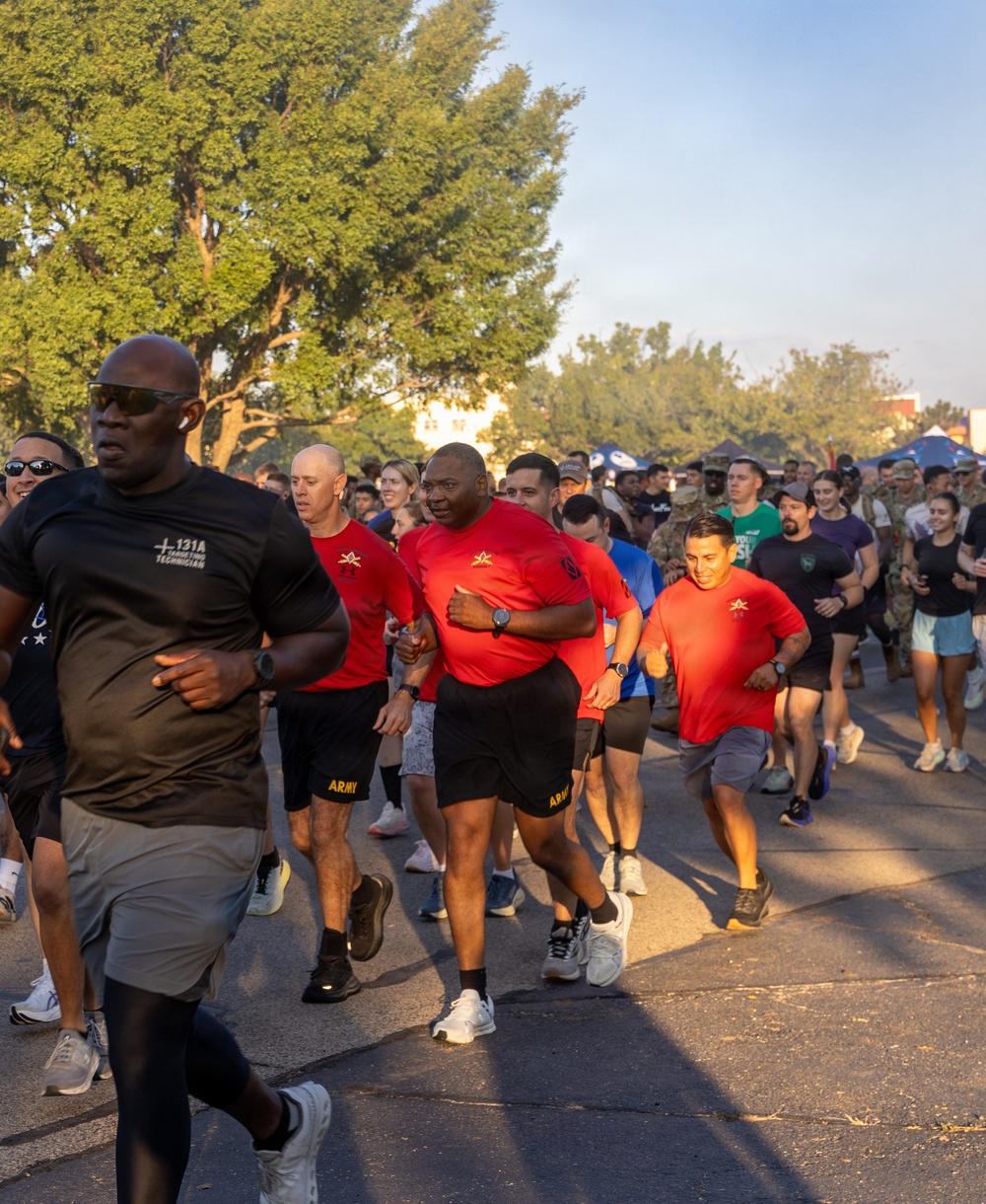 A large group of military personnel and civilians participate in an organized run or fun run event, likely on a military installation. Several participants in the foreground wear matching red shirts with a military insignia and black 'ARMY' shorts, suggesting they are leaders or organizers of the event. The setting appears to be an outdoor paved road lined with trees during early morning or late afternoon based on the warm directional lighting.