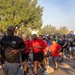 A large group of military personnel and civilians participate in an organized run or fun run event, likely on a military installation. Several participants in the foreground wear matching red shirts with a military insignia and black 'ARMY' shorts, suggesting they are leaders or organizers of the event. The setting appears to be an outdoor paved road lined with trees during early morning or late afternoon based on the warm directional lighting.