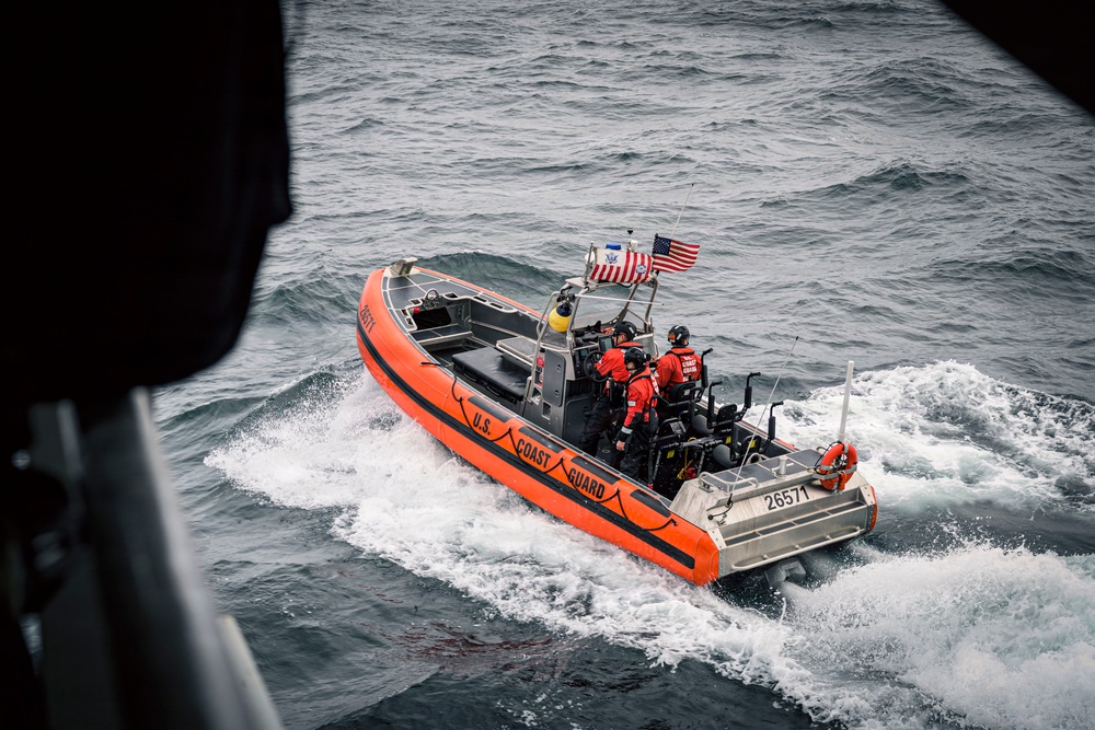A U.S. Coast Guard rigid inflatable boat (RIB) with hull number 26571 is underway on choppy open water. The orange-hulled vessel carries multiple crew members in red survival suits and helmets, with an American flag and what appears to be a Coast Guard ensign flying from the console. The photo appears to be taken from an elevated vantage point, possibly from a larger vessel or aircraft.
