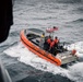 A U.S. Coast Guard rigid inflatable boat (RIB) with hull number 26571 is underway on choppy open water. The orange-hulled vessel carries multiple crew members in red survival suits and helmets, with an American flag and what appears to be a Coast Guard ensign flying from the console. The photo appears to be taken from an elevated vantage point, possibly from a larger vessel or aircraft.
