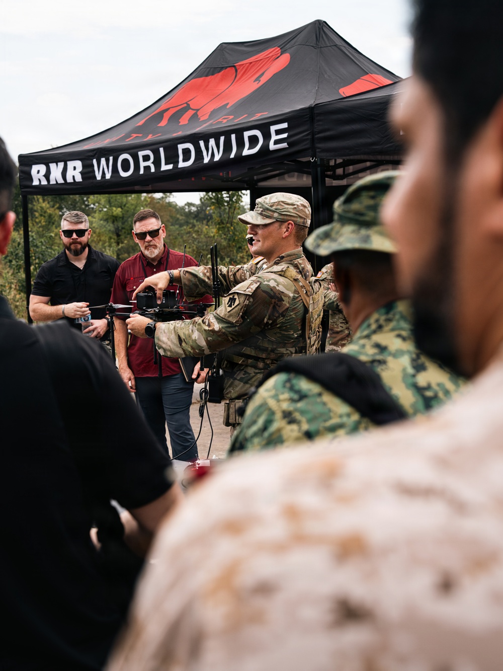 A military demonstration or briefing is taking place outdoors under a black canopy tent branded 'RXR WORLDWIDE'. A soldier in MultiCam/OCP camouflage is presenting or demonstrating a small drone/UAV to a group that includes both military personnel in various camouflage patterns and civilians in dark clothing. The setting appears to be a defense technology expo or field demonstration event.