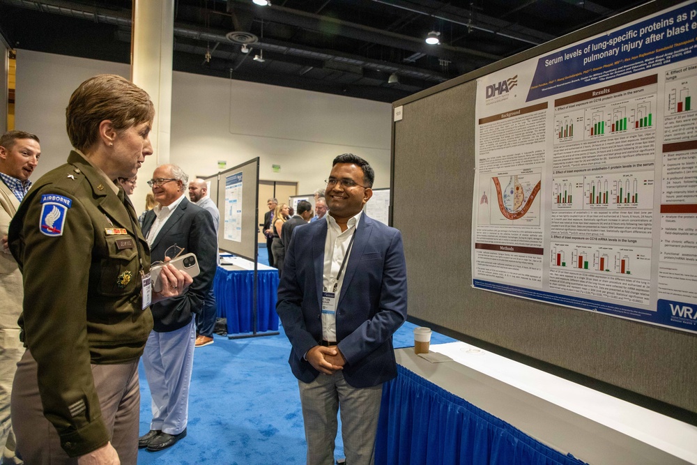 A military officer in Army dress uniform with Airborne patch is engaged in conversation with a civilian researcher at an academic poster session. The research poster, affiliated with DHA (Defense Health Agency) and WRAIR (Walter Reed Army Institute of Research), presents findings on 'Serum levels of lung-specific proteins as pulmonary injury after blast exposure.' The setting appears to be a formal military medical or scientific conference.