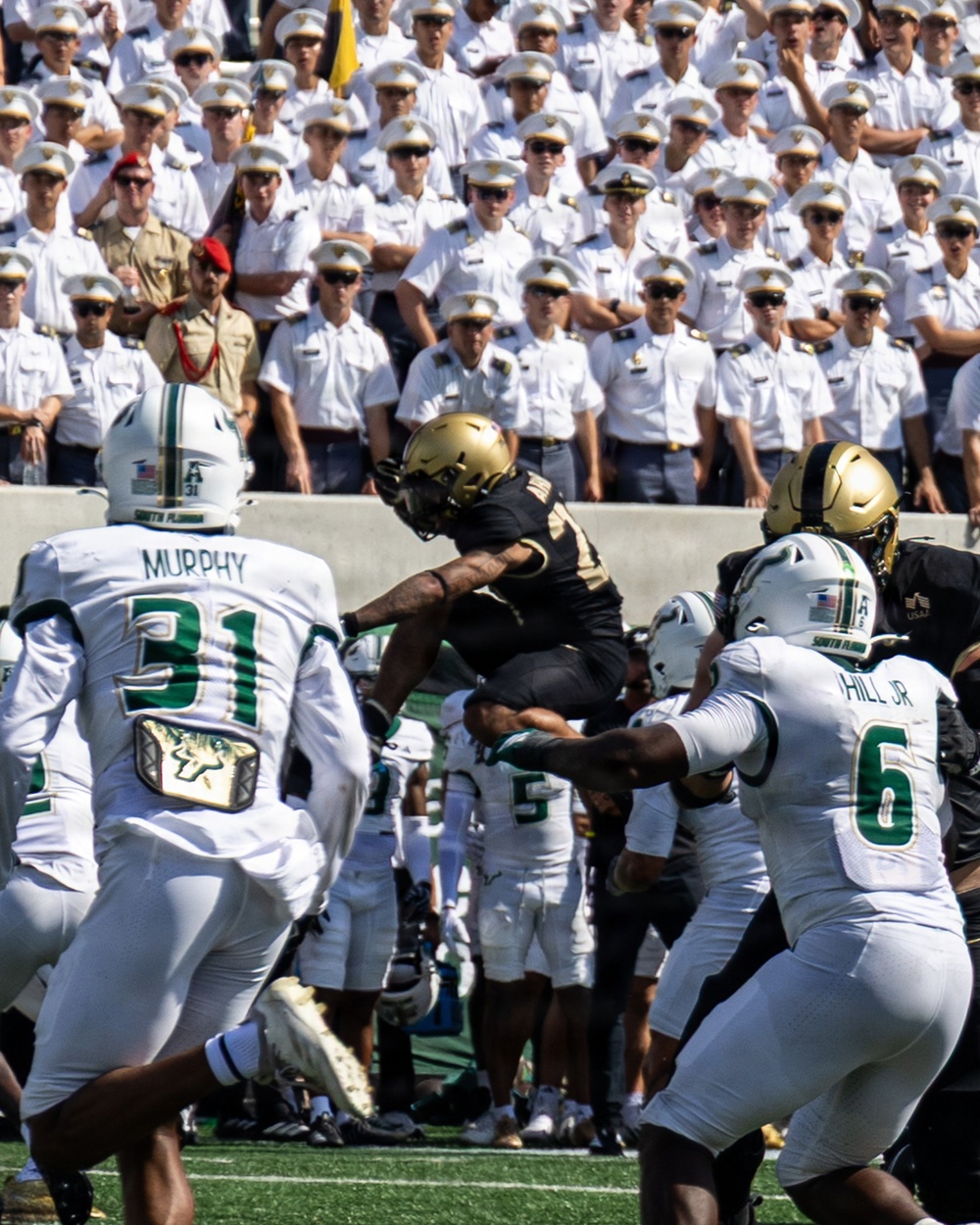 A college football game is in progress, with a player in a black and gold uniform (Army Black Knights) leaping over defenders in white and green uniforms (South Florida Bulls). The background is filled with rows of uniformed military cadets in white dress uniforms, consistent with West Point's Michie Stadium. Two USF defenders are visible — Murphy (#31) and Hill Jr. (#6) — attempting to stop the ball carrier.