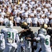 A college football game is in progress, with a player in a black and gold uniform (Army Black Knights) leaping over defenders in white and green uniforms (South Florida Bulls). The background is filled with rows of uniformed military cadets in white dress uniforms, consistent with West Point's Michie Stadium. Two USF defenders are visible — Murphy (#31) and Hill Jr. (#6) — attempting to stop the ball carrier.