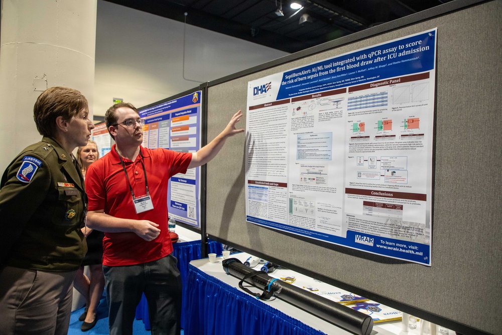 A man in a red polo shirt with a conference lanyard is presenting a research poster to a woman in a U.S. Army military uniform with Airborne insignia at what appears to be a scientific or military research conference. The poster is titled 'SeptiBurnAlert: AI/ML tool integrated with qPCR assay to score the risk of burn sepsis from the first blood draw after ICU admission' and is affiliated with DHA (Defense Health Agency) and WRAIR. A third person is partially visible in the background.
